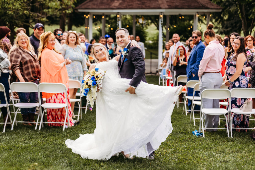 A happy couple in wedding attire is posing playfully amid guests during an outdoor wedding ceremony. The mood is joyful, with vibrant colors and smiles all around.