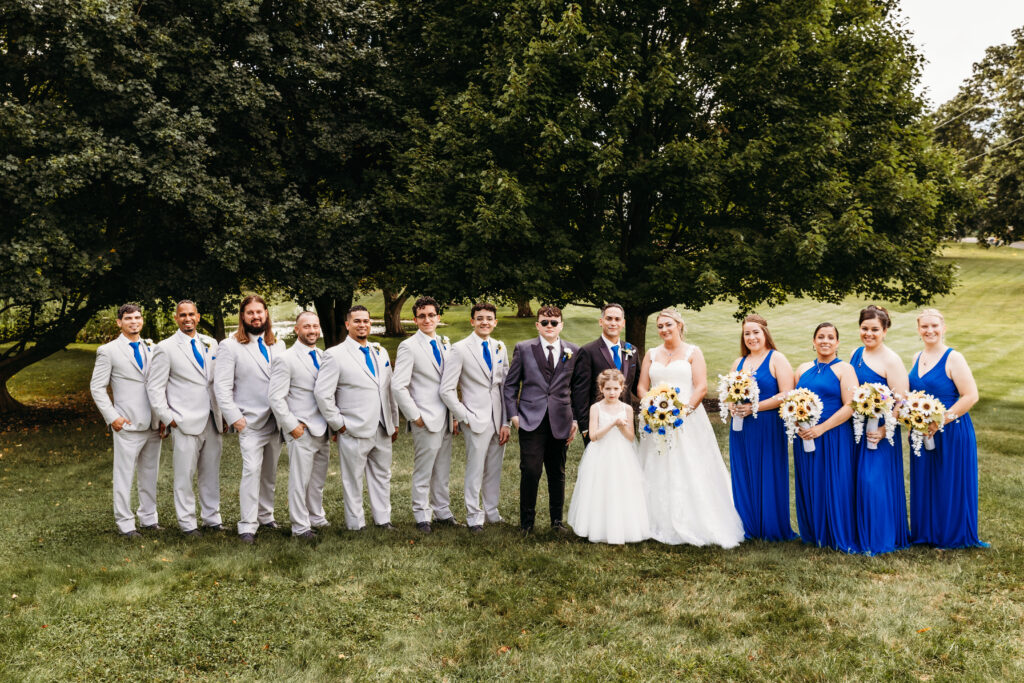 A wedding party poses outdoors, featuring people in gray suits with blue ties, two in white and a child, alongside others in blue dresses holding bouquets.