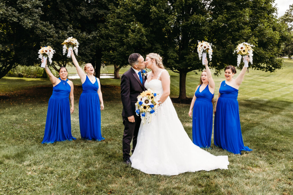 A wedding scene with a person in a white dress kissing another person while four people in blue dresses cheer with bouquets in a park.