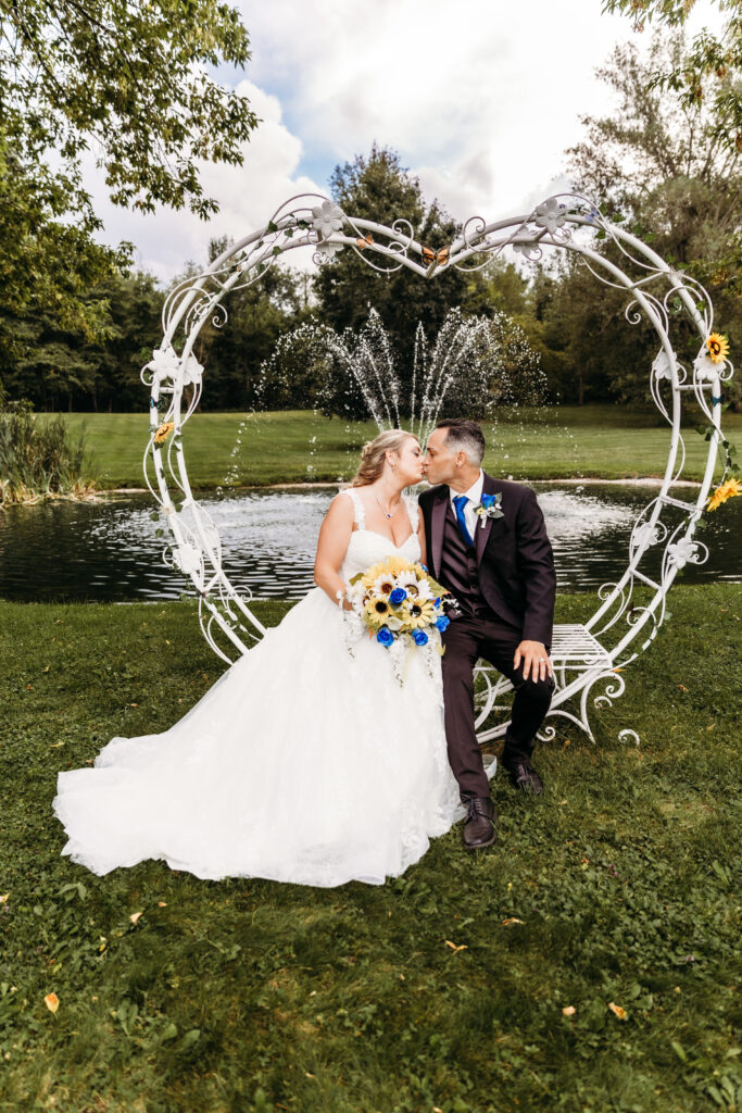 Two people in wedding attire kiss under a decorative heart-shaped arch by a pond with a fountain. The setting is serene with greenery.