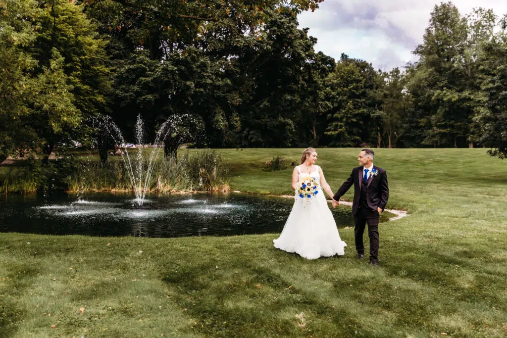 Two people in wedding attire hold hands near a pond with a fountain in a lush green park, symbolizing a special matrimonial moment.