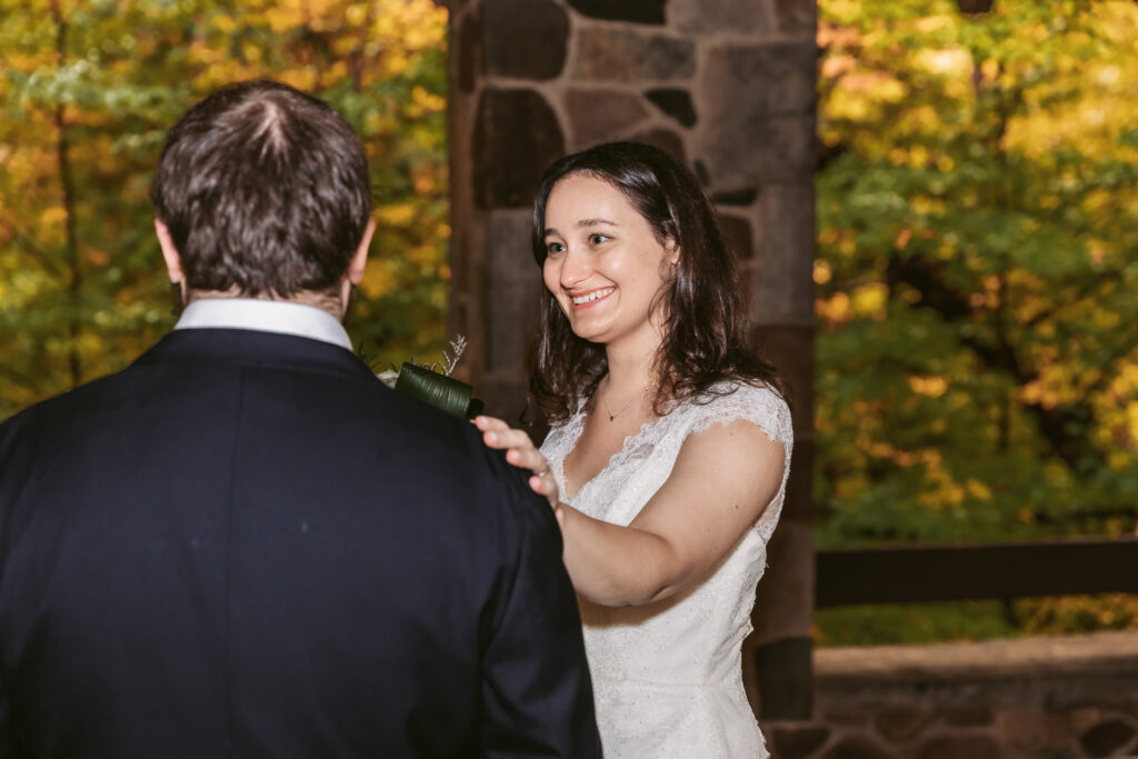 A person in a white dress smiles at another person outdoors, with autumn foliage and stone structure in the background.