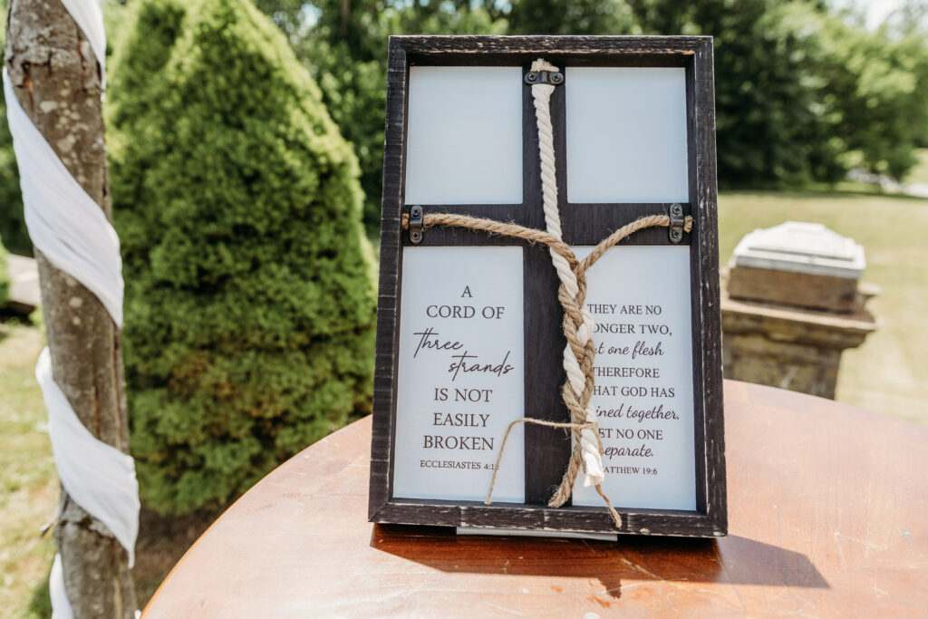 A rustic decorative frame with biblical quotes about marriage, entwined with a cord, is displayed outdoors, possibly at a wedding venue.