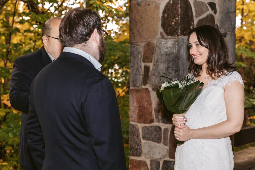 Three people in formal attire, one holding flowers, stand near a stone wall, surrounded by lush greenery, during a likely outdoor event.