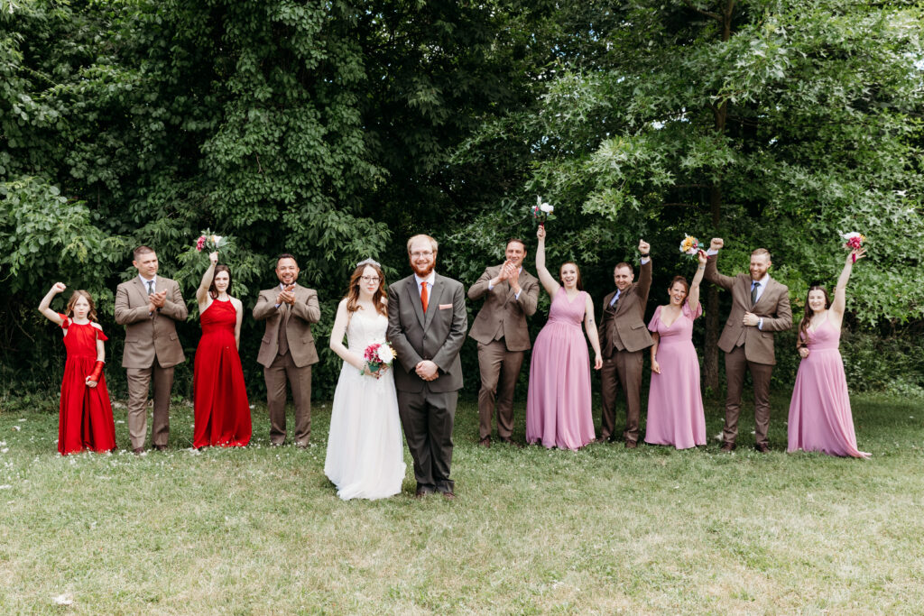 A wedding party outdoors, with a happy couple in the center. People in suits and dresses are posing with bouquets, some with raised arms.
