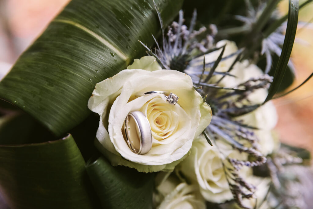 Close-up of a white rose bouquet with green leaves, featuring silver rings and a diamond ring nestled among the petals and greenery.