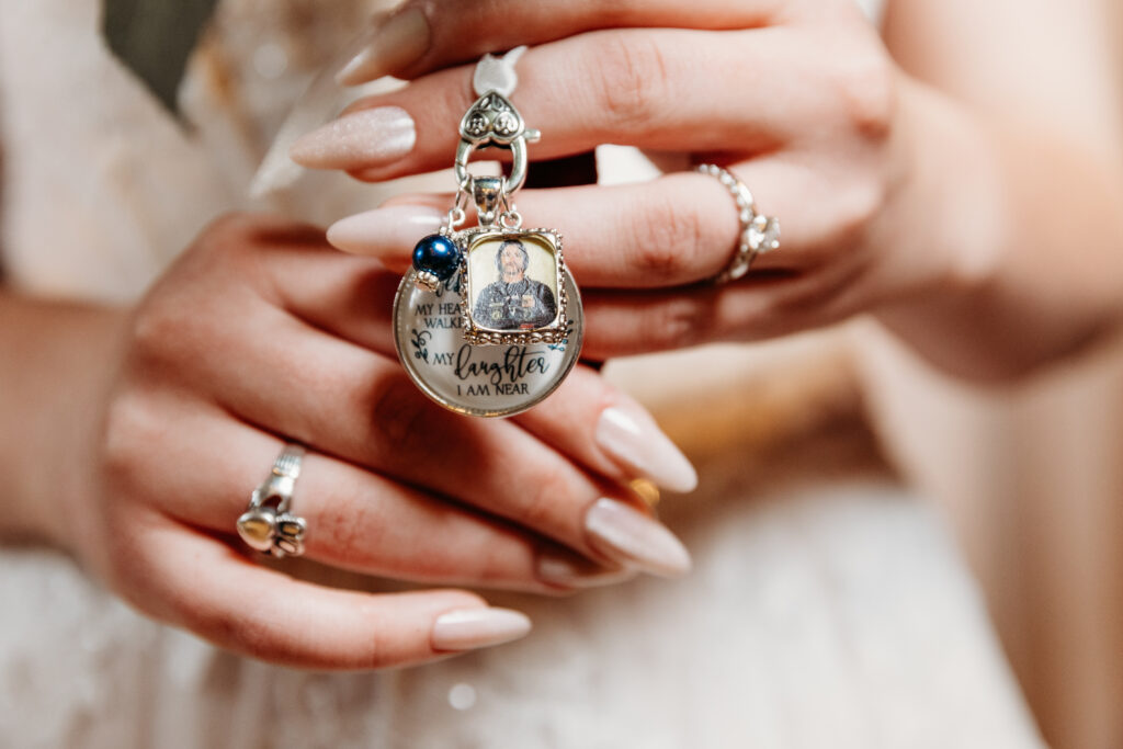 A person holds a charm with a photo and sentimental text, suggesting a memorial or tribute, perhaps for a wedding. Rings and well-manicured nails are visible.