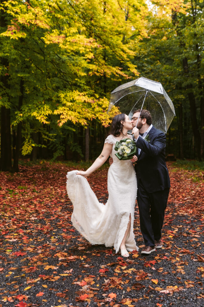 Two people in formal attire share a kiss under a clear umbrella amidst vibrant autumn foliage on a leaf-strewn path.