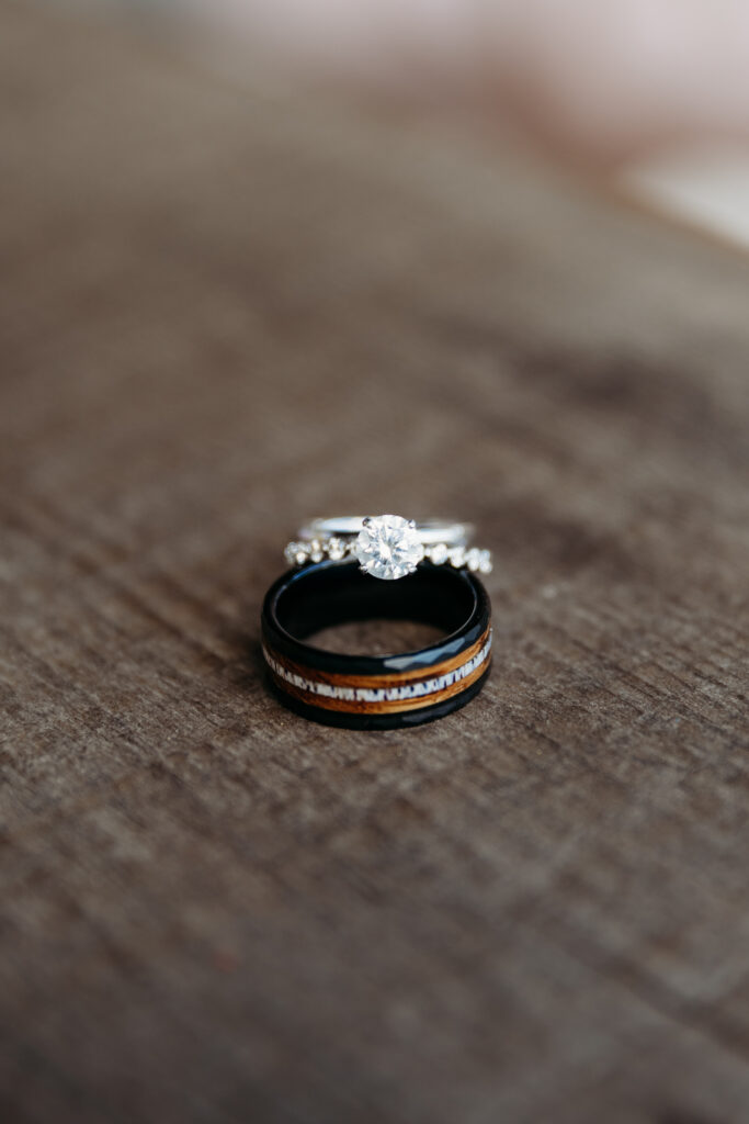 Two wedding rings on a textured surface; one with a large diamond, the other black with a wood inlay. The focus is on the sparkling gemstone.
