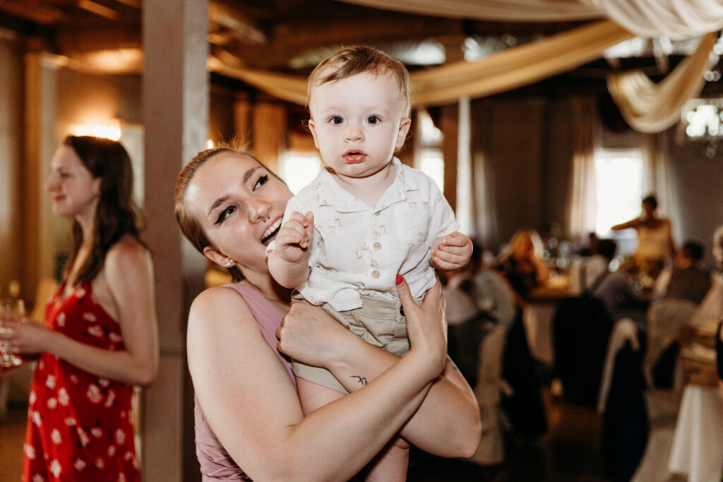 A person holding a toddler smiles at the camera amidst a festively decorated indoor space with guests, suggestive of a celebration or gathering.