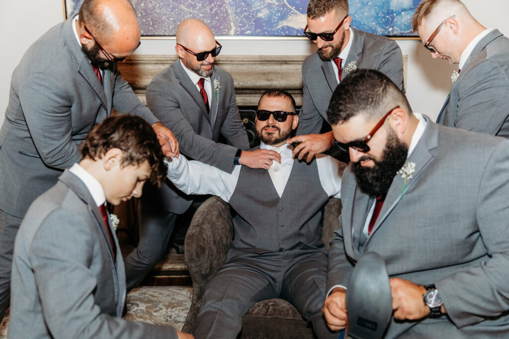A group of people in suits, wearing sunglasses, are helping a seated person adjust their tie in an indoor setting.