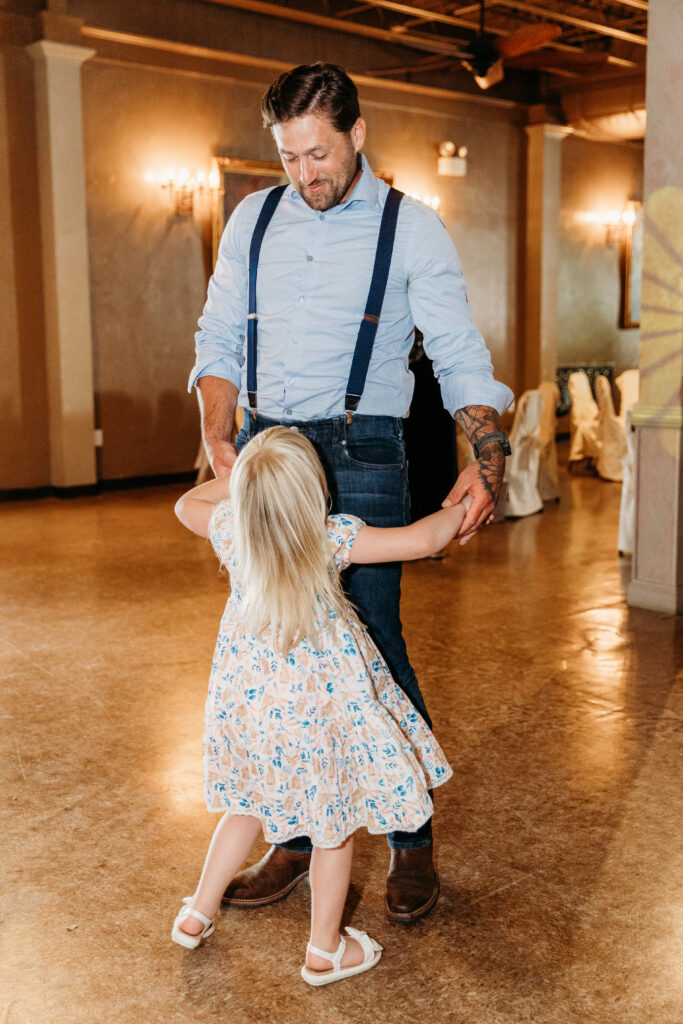 A person with a beard and tattoos is joyfully dancing with a young girl in a flower-patterned dress in a warmly lit indoor environment.