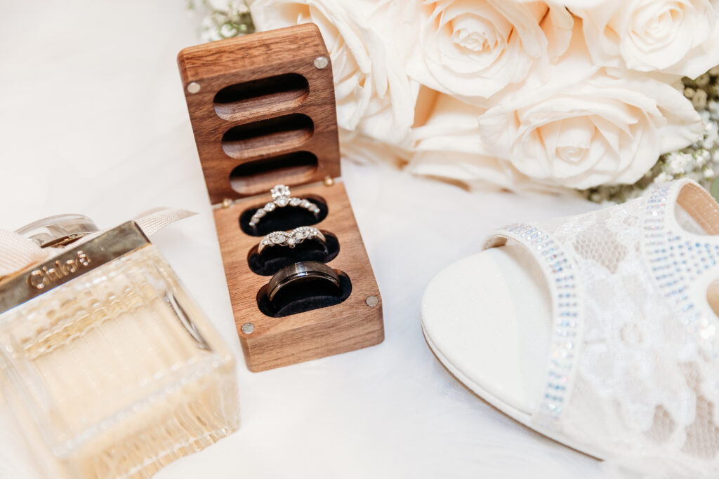 A wedding themed photograph featuring rings on a wooden stand, cream roses, bridal shoes, and a bottle of perfume on a white background.