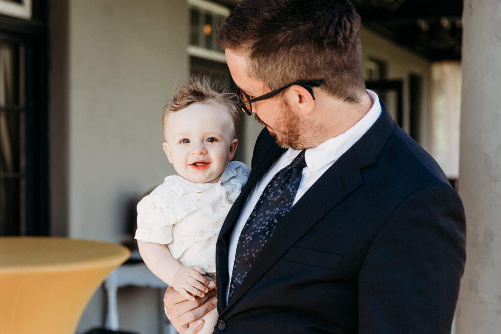 A person in a suit holds a cheerful baby. They share a joyful moment outside with soft lighting and a neutral-colored background.