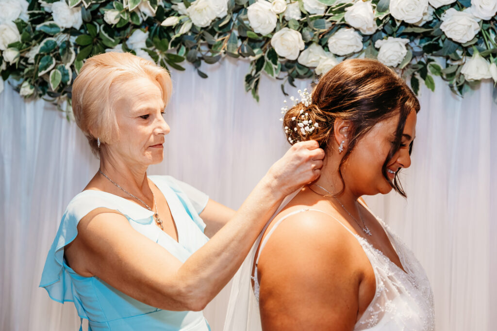 Two people, one adjusting the other's hair, in front of a backdrop adorned with white roses and greenery. No recognizable landmarks or historical buildings present.