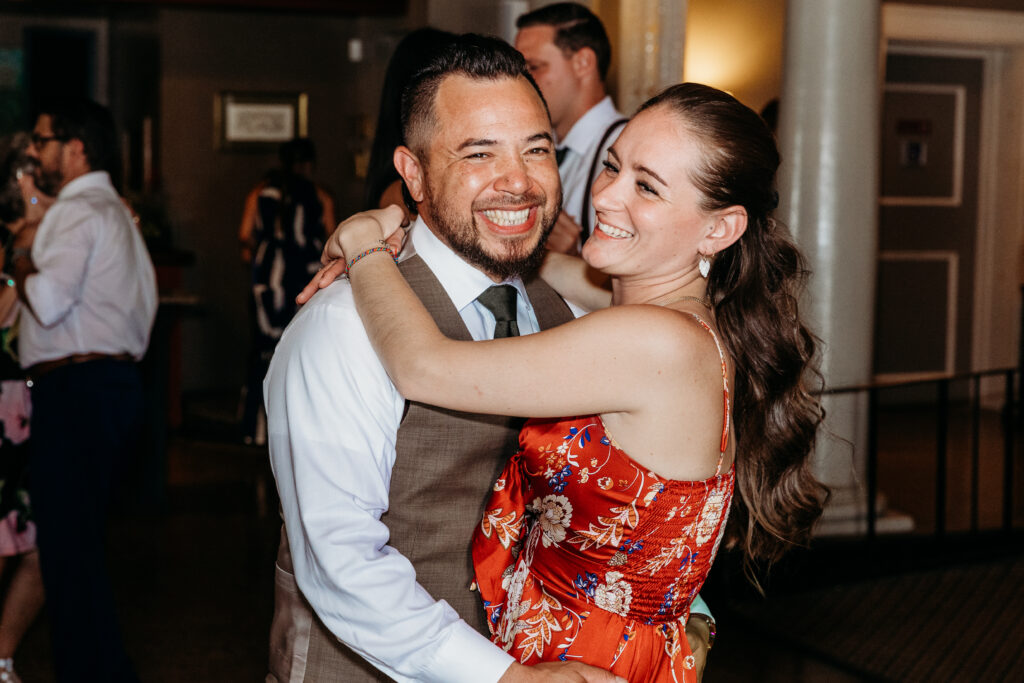 Two people are joyfully dancing together at an event. The person in the foreground wears a gray vest, while the other has a floral dress. They're smiling brightly.
