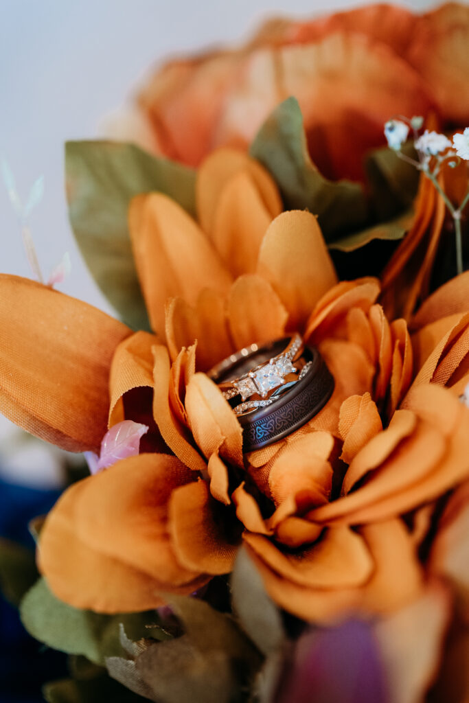 Two rings nestled in vibrant orange flowers, showcasing a sparkling diamond against intricate patterns, symbolizing elegance and commitment amidst a floral backdrop.