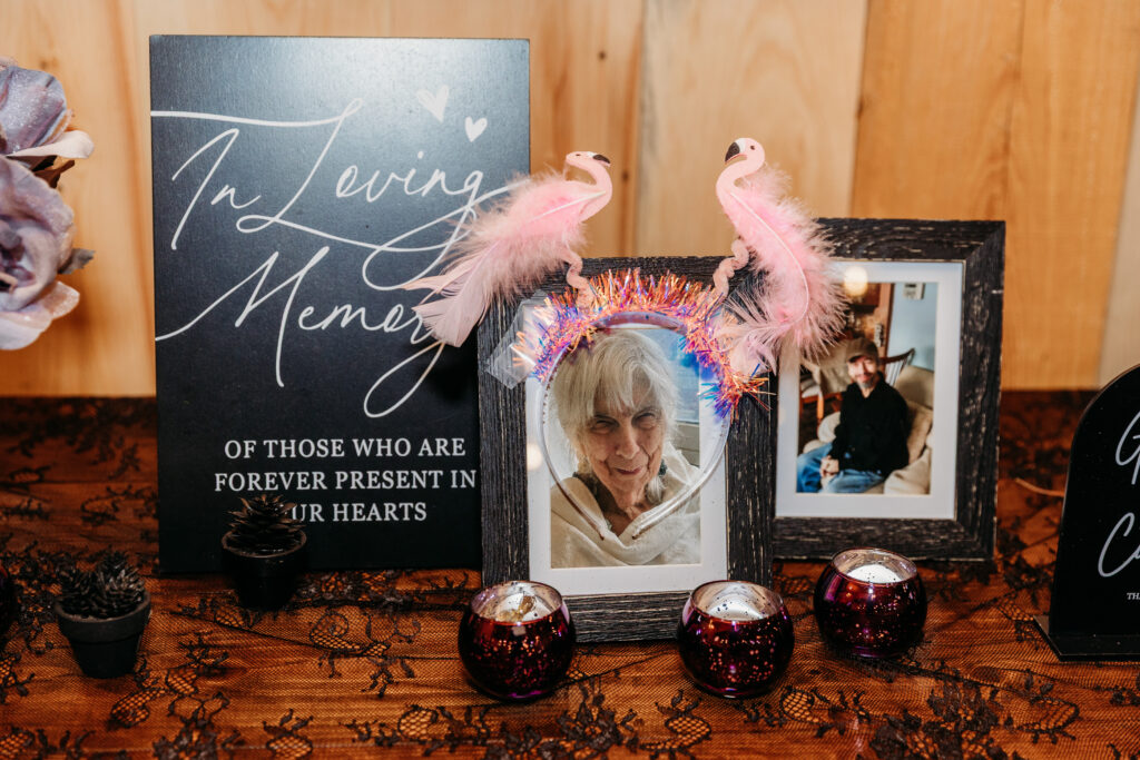 A memorial display on a wooden surface featuring an “In Loving Memory” sign, framed photos, pink feather decorations, and small candle holders.