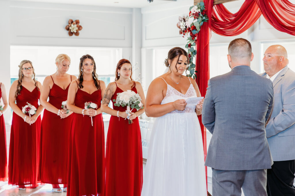 A wedding ceremony with a person in white reading vows and five individuals in red dresses holding flowers beside a person in a suit.