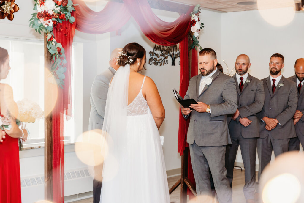 A wedding ceremony with a person in a grey suit reading, adorned arch with flowers, and attendants in red dresses and grey suits.