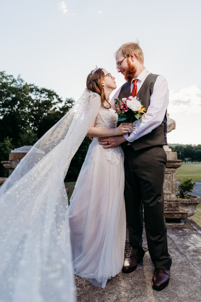 Two people in wedding attire are intimately smiling at each other, likely a bride and groom, outdoors with a clear sky in the background.