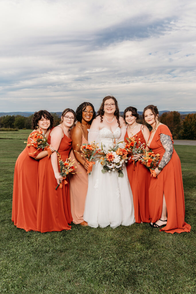 A bride in a white wedding dress stands with five bridesmaids in orange gowns on a grassy field outdoors, all holding bouquets.