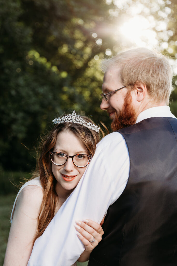 Two people in wedding attire share a joyful moment. The person in the foreground wears glasses and a tiara, embracing their partner, in soft sunlight.
