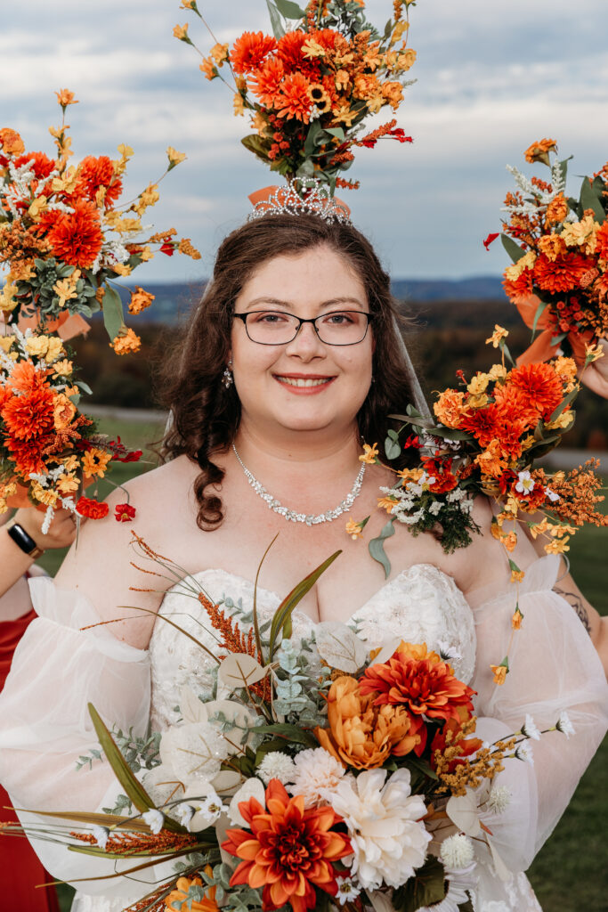 Bride wearing glasses in a white wedding dress with a floral crown and a bouquet of orange and white flowers, outdoors.