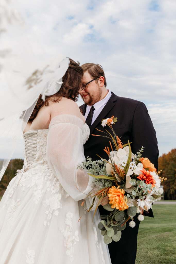 A bride and groom kiss outdoors at a wedding, the bride in a white lace gown with a veil and the groom in a dark suit, holding a bouquet of orange and white flowers.