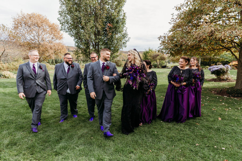 A wedding party strolling across a grassy park in fall, with men in gray suits and purple accents and women in purple dresses.