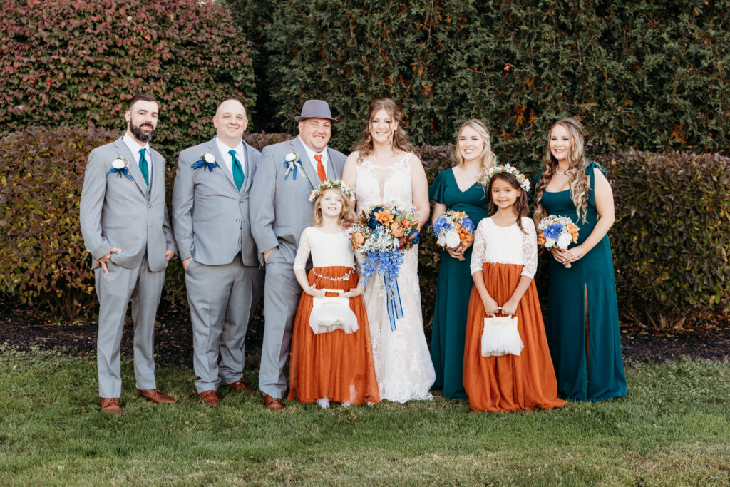 A group of people dressed formally in front of shrubbery, holding bouquets, posing for a photograph on grass, includes children in festive attire.
