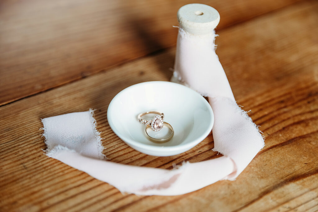 A white ceramic dish with a pair of rings rests on a wooden surface, partially wrapped in a soft pink fabric with frayed edges.