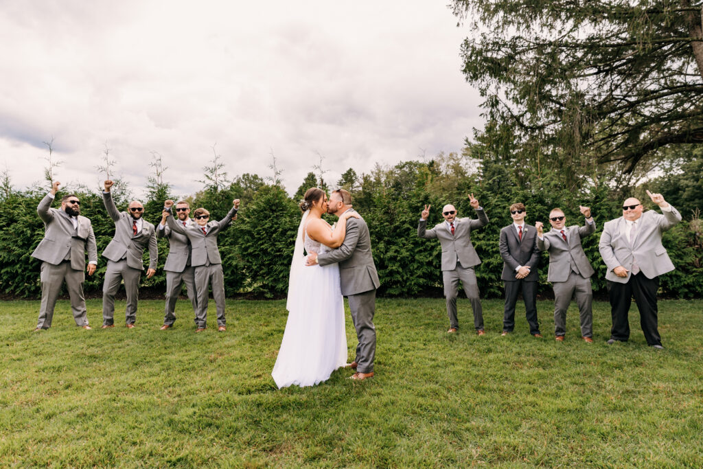 A couple kisses outdoors in front of eight joyful people in gray suits, celebrating on a grassy area with trees in the background.