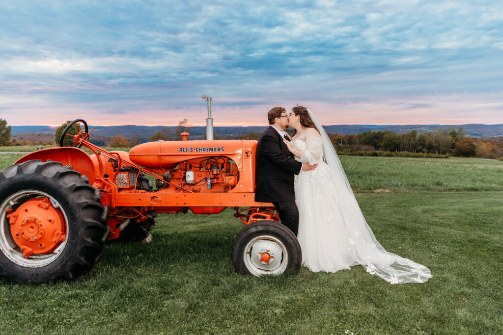 Bride and groom kiss beside a vintage orange tractor on a grassy field under a blue sky.