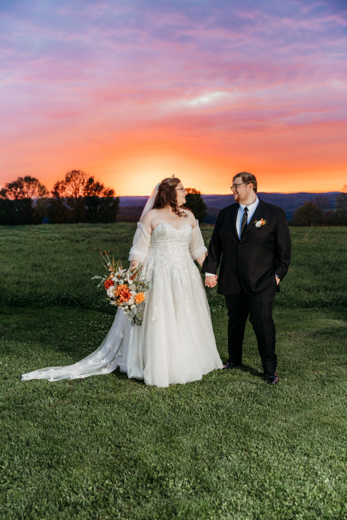 A bride in a white wedding gown with a long veil and an orange bouquet stands hand in hand with a groom in a black suit on a grassy field at sunset.