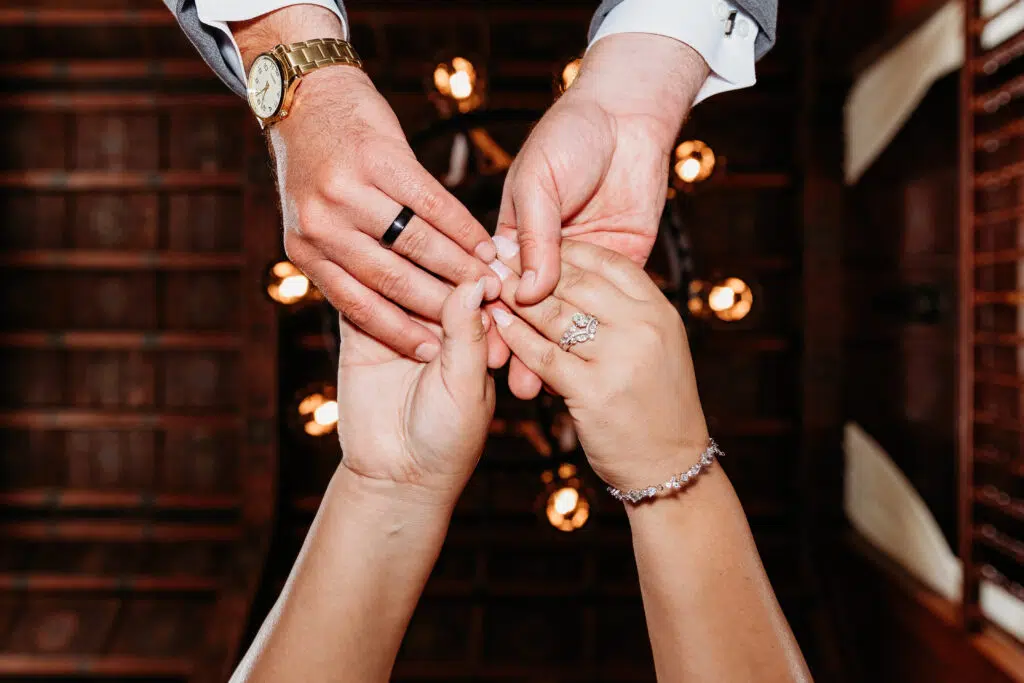 Two people exchange rings, likely during a wedding ceremony. Warm lights glow in the background, complementing the intimate and celebratory moment.