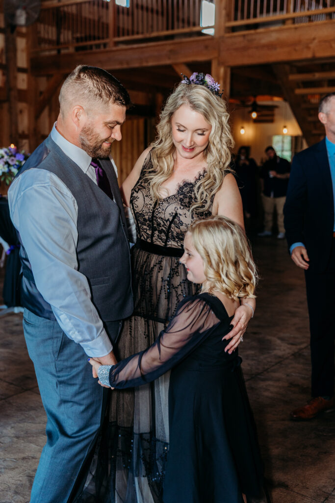 Man in a vest and tie, a woman in a lace dress, and a young girl in a blue dress hold hands and smile at each other in a wooden indoor venue.