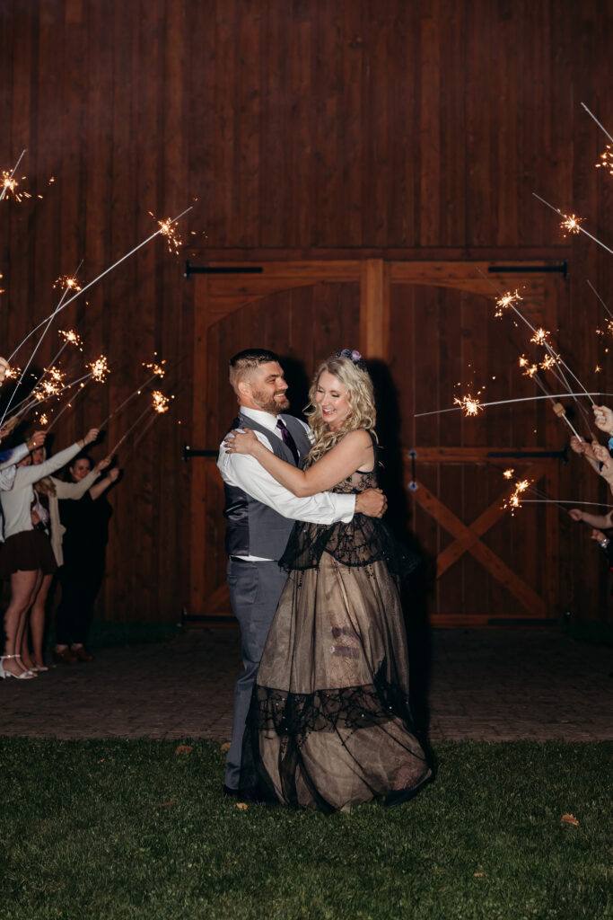 A couple in formal attire dances and embraces in front of a wooden doorway at night, surrounded by guests waving sparklers.