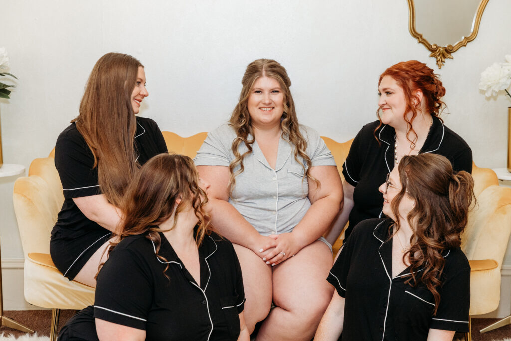 Five women sit on a beige sofa with orange cushions in a bright living room, smiling at the camera.