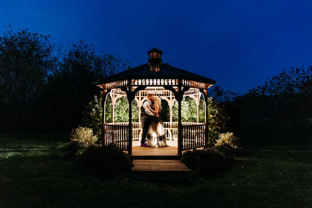 A couple in wedding attire embraces inside a warmly lit wooden gazebo at night.