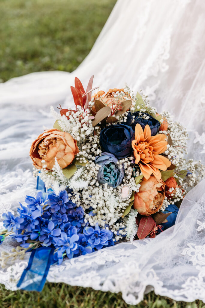 A vibrant bouquet with orange and blue flowers lies on a lacy white fabric, set on green grass outdoors.