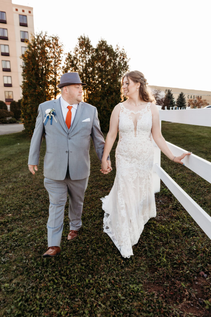 Two people in formal attire walk hand-in-hand near a hotel, with trees and grass in the background. A casino building is visible.