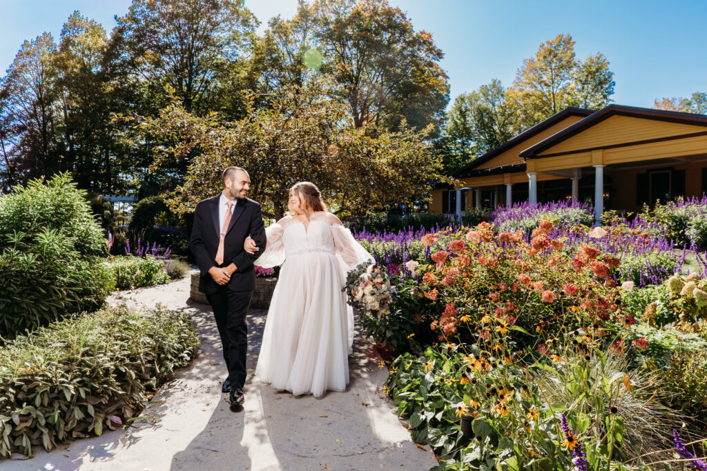 Bride in a white wedding dress and groom in a dark suit walk together along a gravel garden path lined with colorful flowers, with a building in the background.