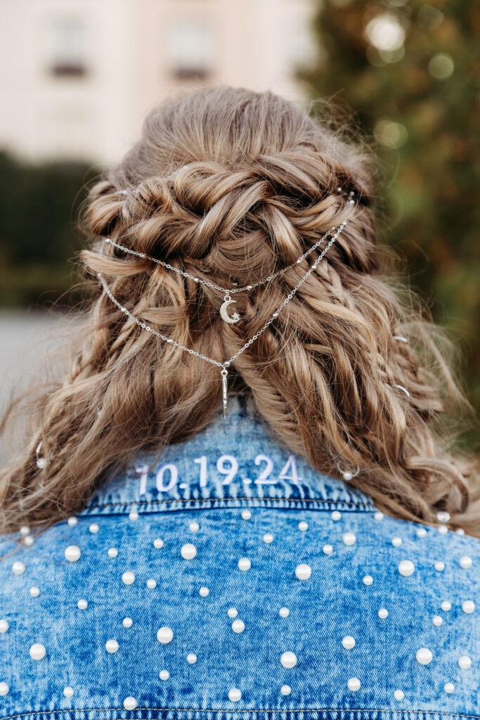 Person with intricate braided hairstyle and decorative chain in hair, wearing a denim jacket with pearls and a date written on it.