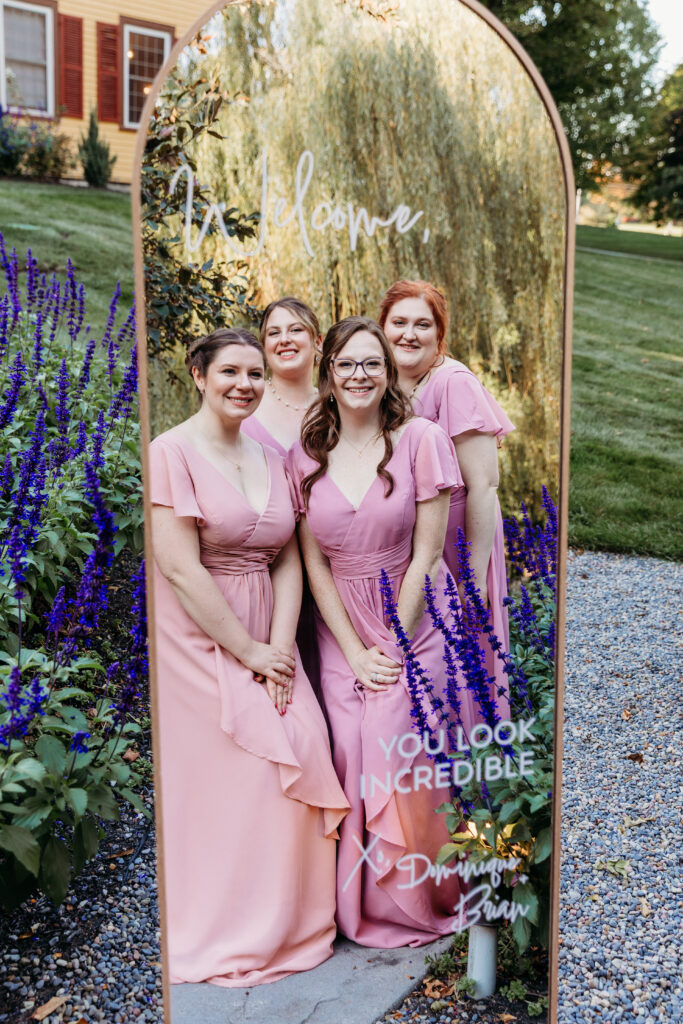 Four bridesmaids in pink dresses pose in front of a gold arched mirror backdrop with purple flowers in a garden.