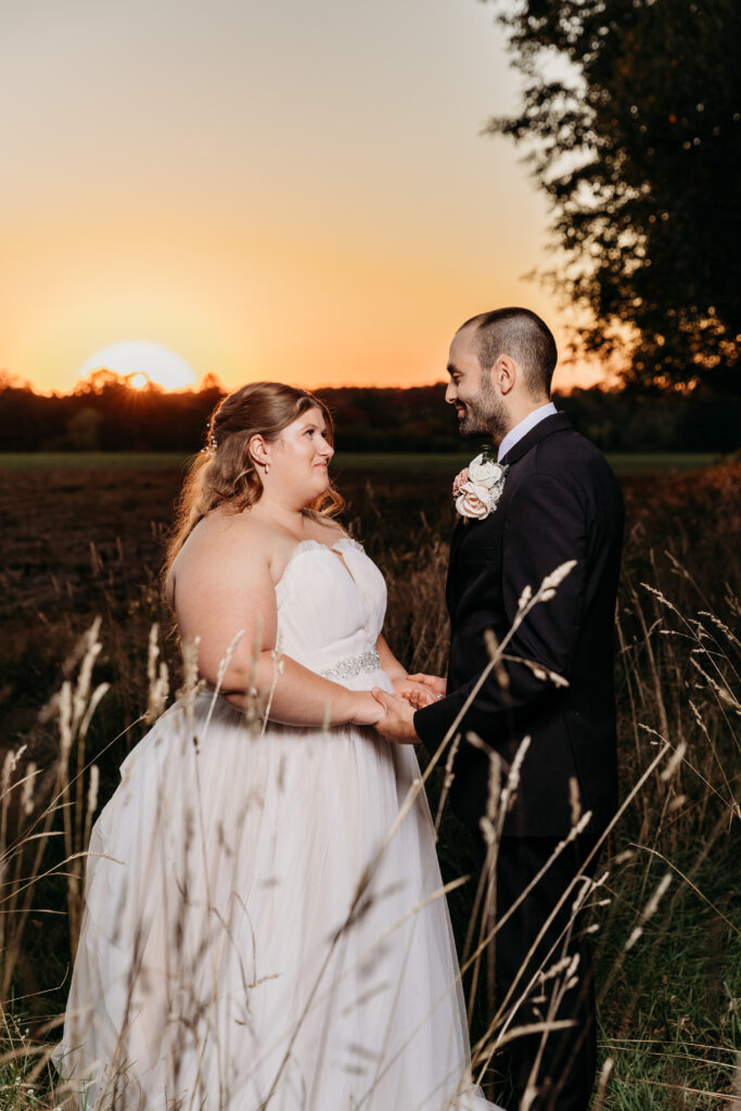 A bride in a white wedding gown and a groom in a dark suit stand in a grassy field at sunset, holding hands and smiling at each other.