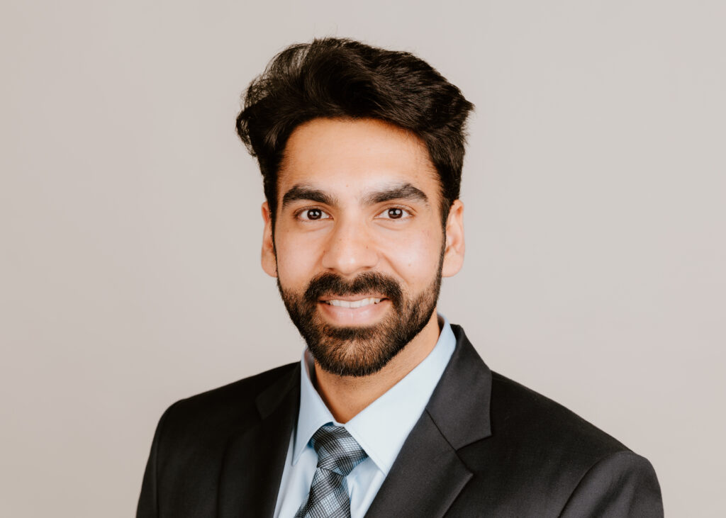 A person with dark hair and a beard smiles warmly at the camera, wearing a dark suit, tie, and light blue shirt against a neutral background.