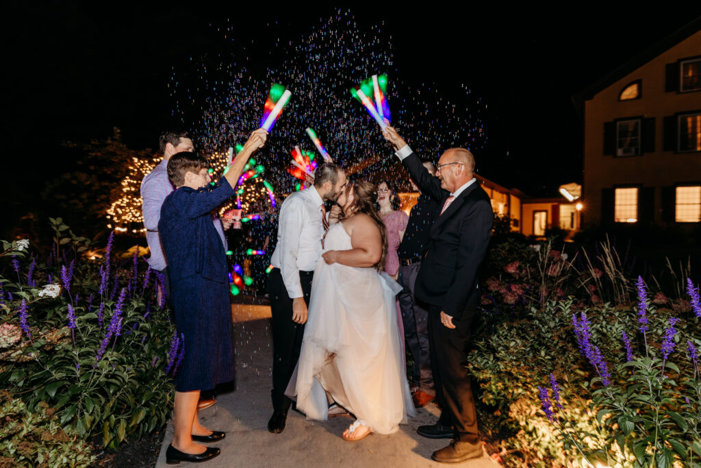 Bride and groom celebrate their wedding at night as guests shower them with rainbow confetti in a flower-filled garden.