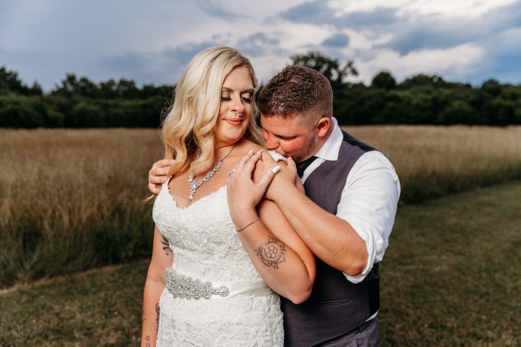 A bride in a white wedding dress and a groom in a gray vest embrace in a grassy field as he kisses her shoulder.