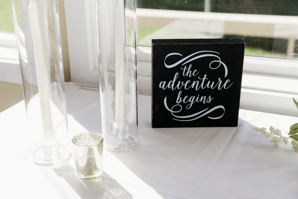 Decorative black sign reading 'the adventure begins' sits on a white table beside glass vases and a small candle holder near a window.